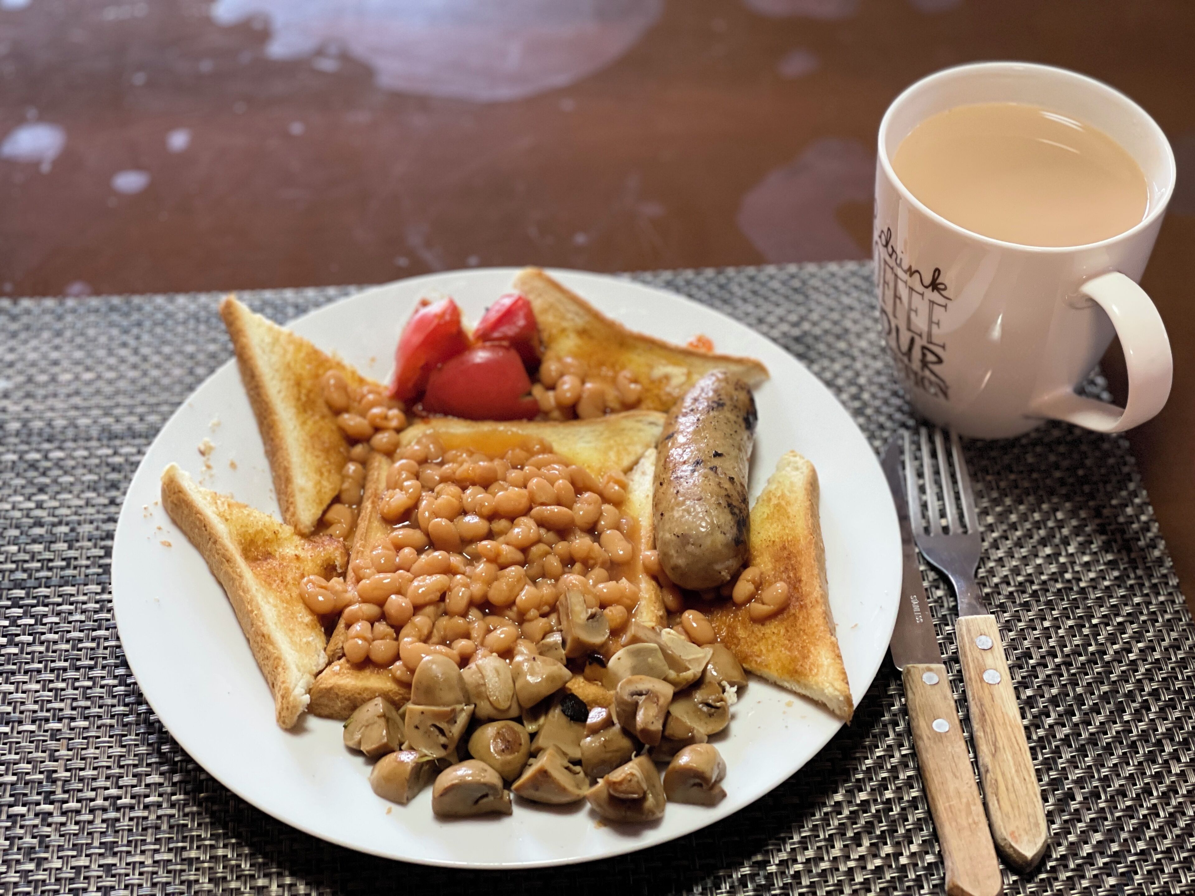 Plat de menjar amb fesols, bolets i una salsitxa, acompanyat d'una tassa de cafè i un garfor sobre un mantell de taula trenat.