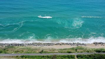 An aerial view of a beach with waves crashing onto the shore, a road running parallel to the beach, and a dense green area in the foreground.