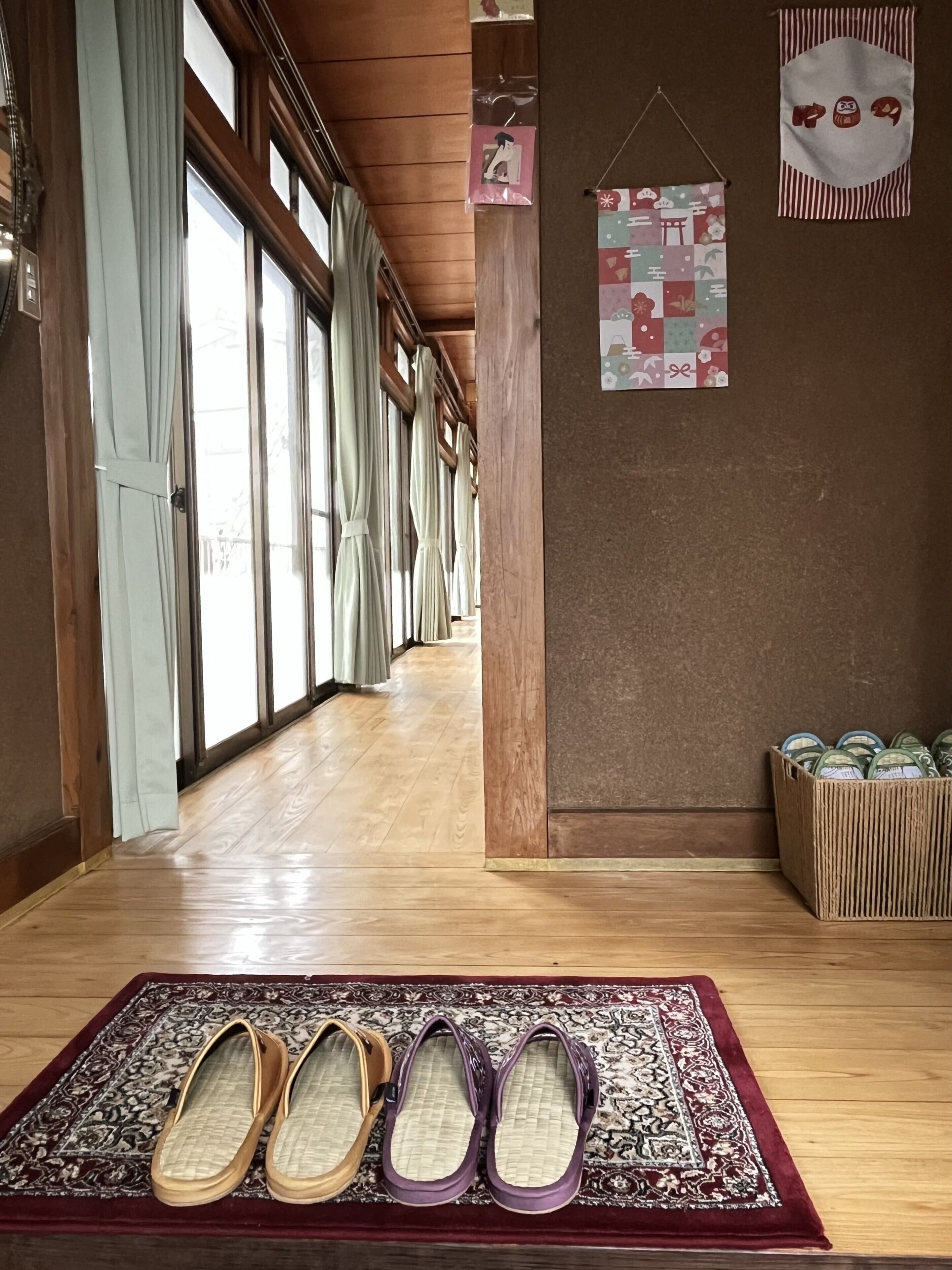 A hallway with wooden floors, large windows, and a patterned rug with slippers.