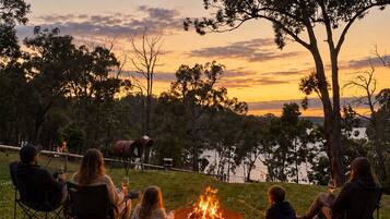 Eine Gruppe von Menschen sitzt an einem Lagerfeuer in einer Outdoor-Umgebung, im Hintergrund sind Bäume und ein Gewässer zu sehen.