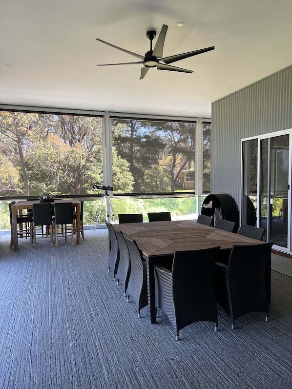 A covered patio with a dining table and chairs, a ceiling fan, and a view of trees through large windows.