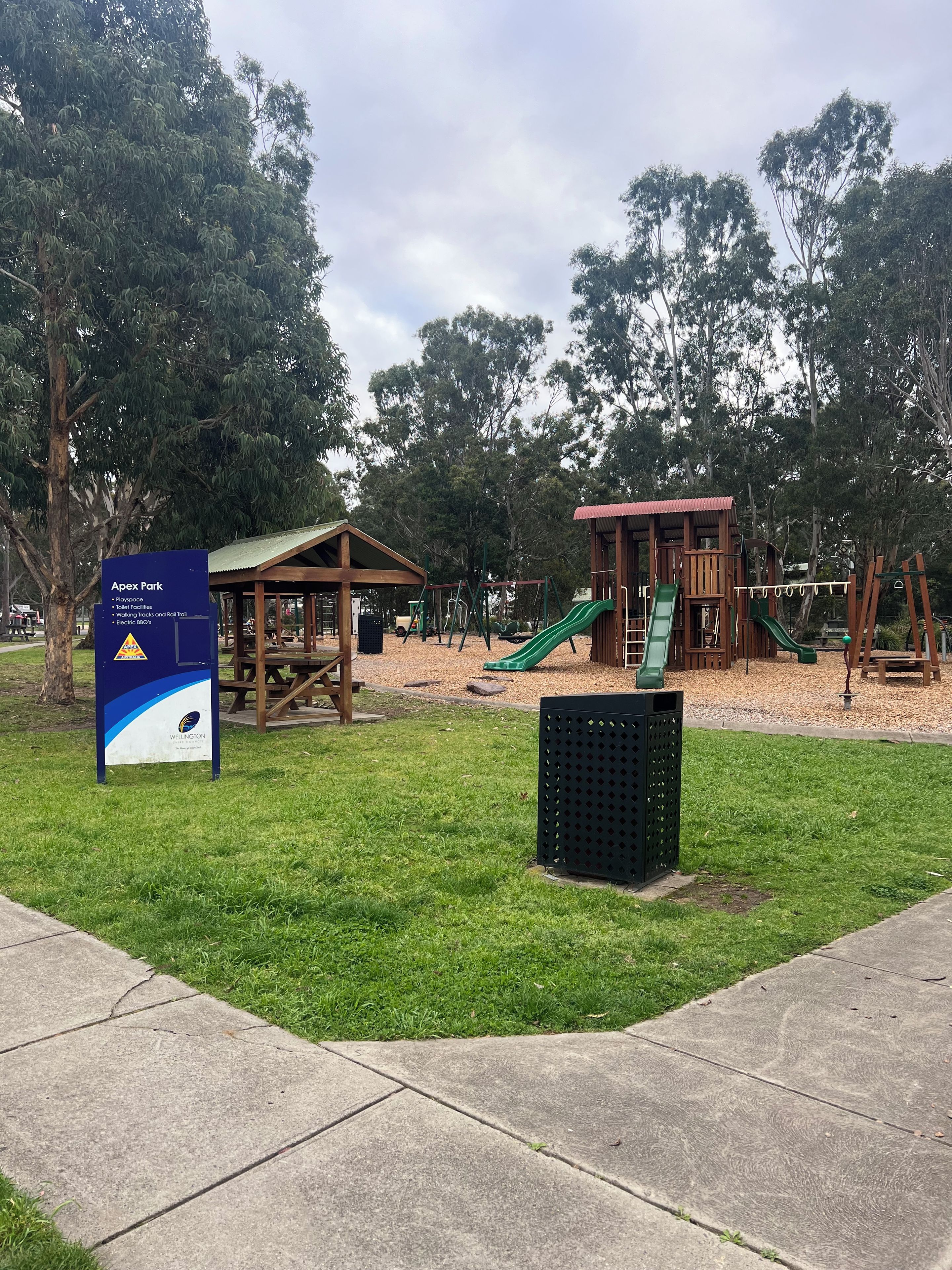 Ein Park mit einem Spielplatz, einer Holzlaube und einem Hinweisschild.