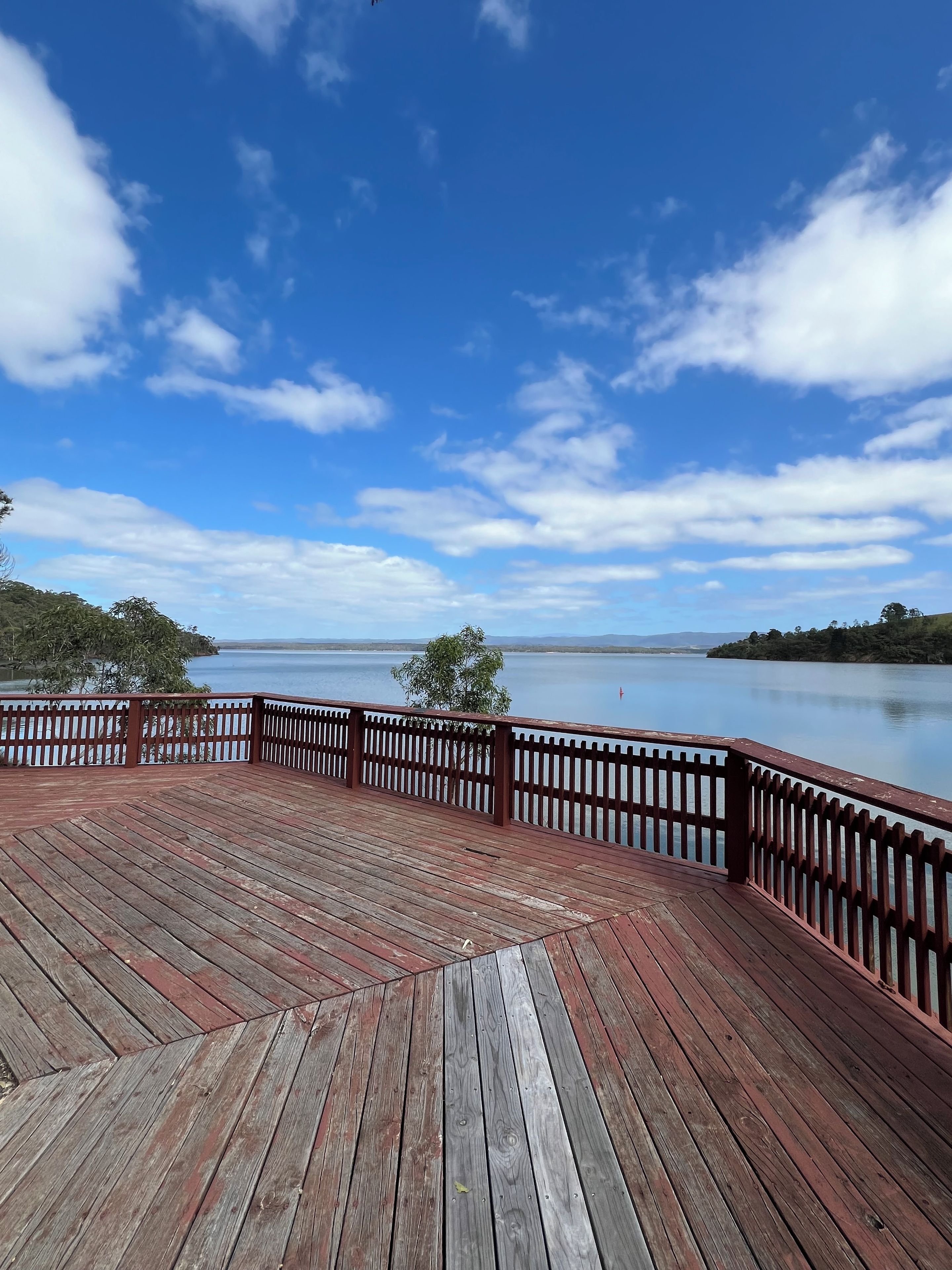 A wooden deck with a railing overlooking a calm body of water and distant hills.