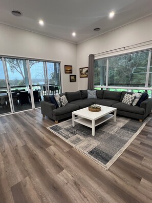 A modern living room with a sectional sofa, a white coffee table, and a large window offering a view of the outdoors. - Lakeside Coongulla - Waterfront Holiday Home - Sleeps 10 + (Coongulla)