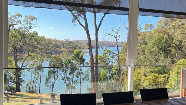 Ein Zimmer mit großem Fenster mit Blick auf einen See und Bäume. Ein Holztisch mit Stühlen und ein Korbsessel sind vorhanden.
