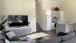 Ein modernes Wohnzimmer mit einem grauen Sofa, einem Flachbildfernseher und einem kleinen weißen Couchtisch. Im Hintergrund ist eine Küche mit weißen Schränken und einer grauen Arbeitsplatte erkennbar.
