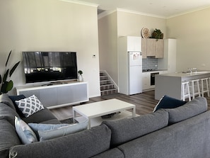 A modern living room with a grey sofa, a flat-screen TV, and a small white coffee table. A kitchen with white cabinets and a grey countertop is visible in the background.