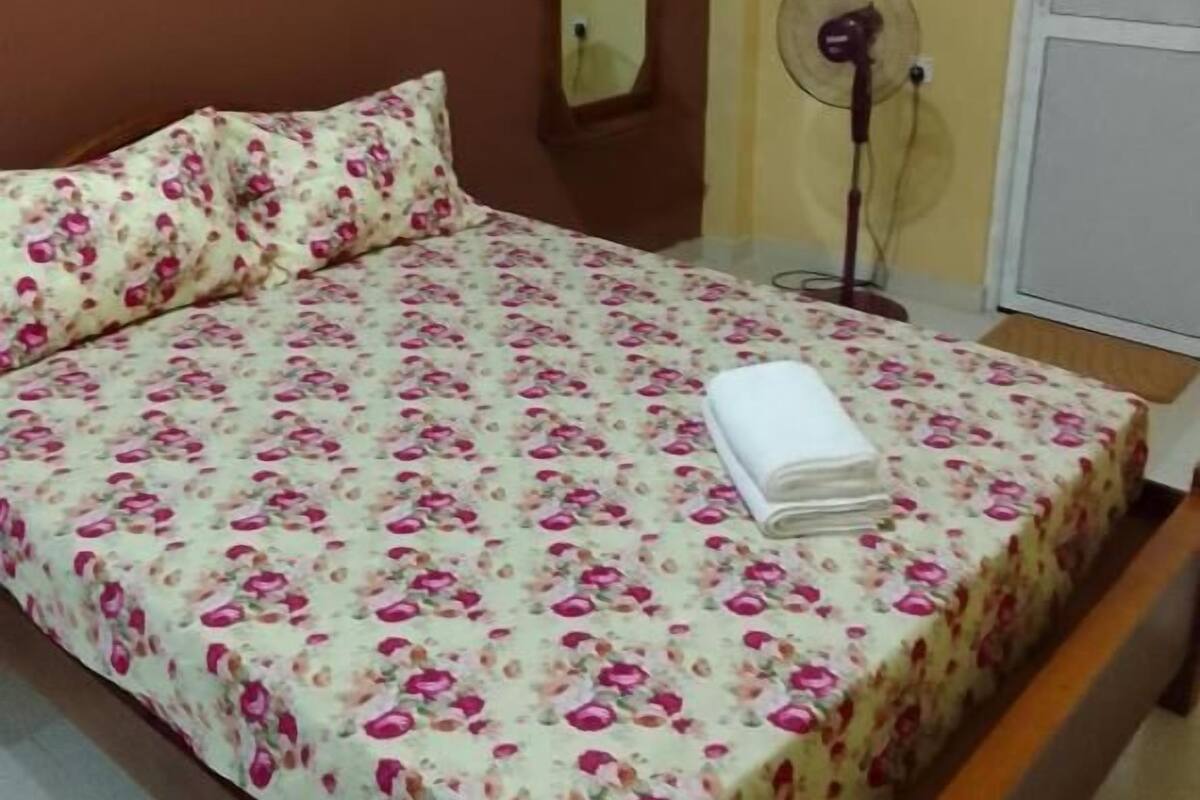 A bed with a floral bedspread, a bedside table with a rolled-up white towel, a standing fan, and a mirror on the wall.