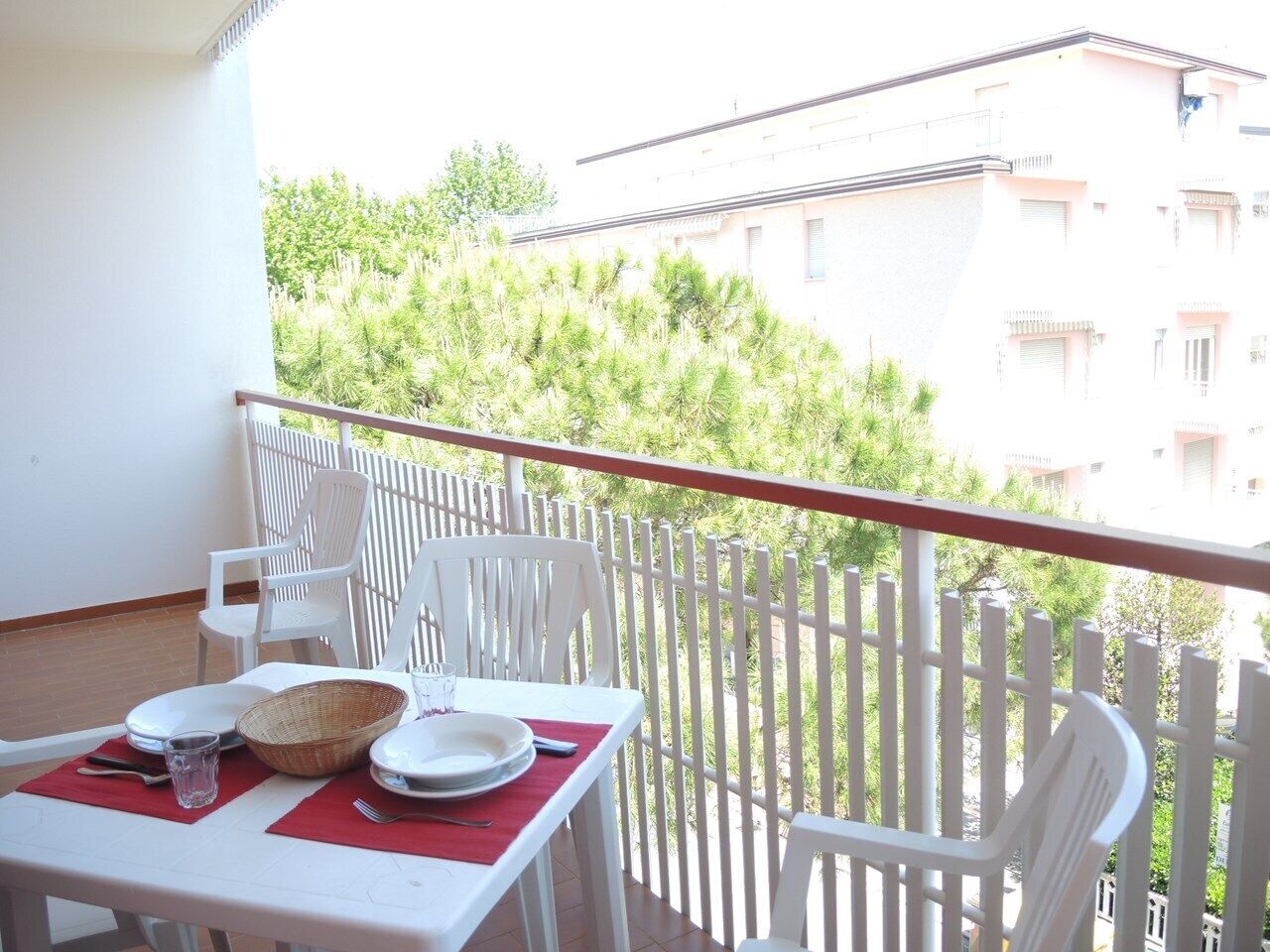 A balcony with a table set for two, white chairs, and a view of greenery and buildings in the background.