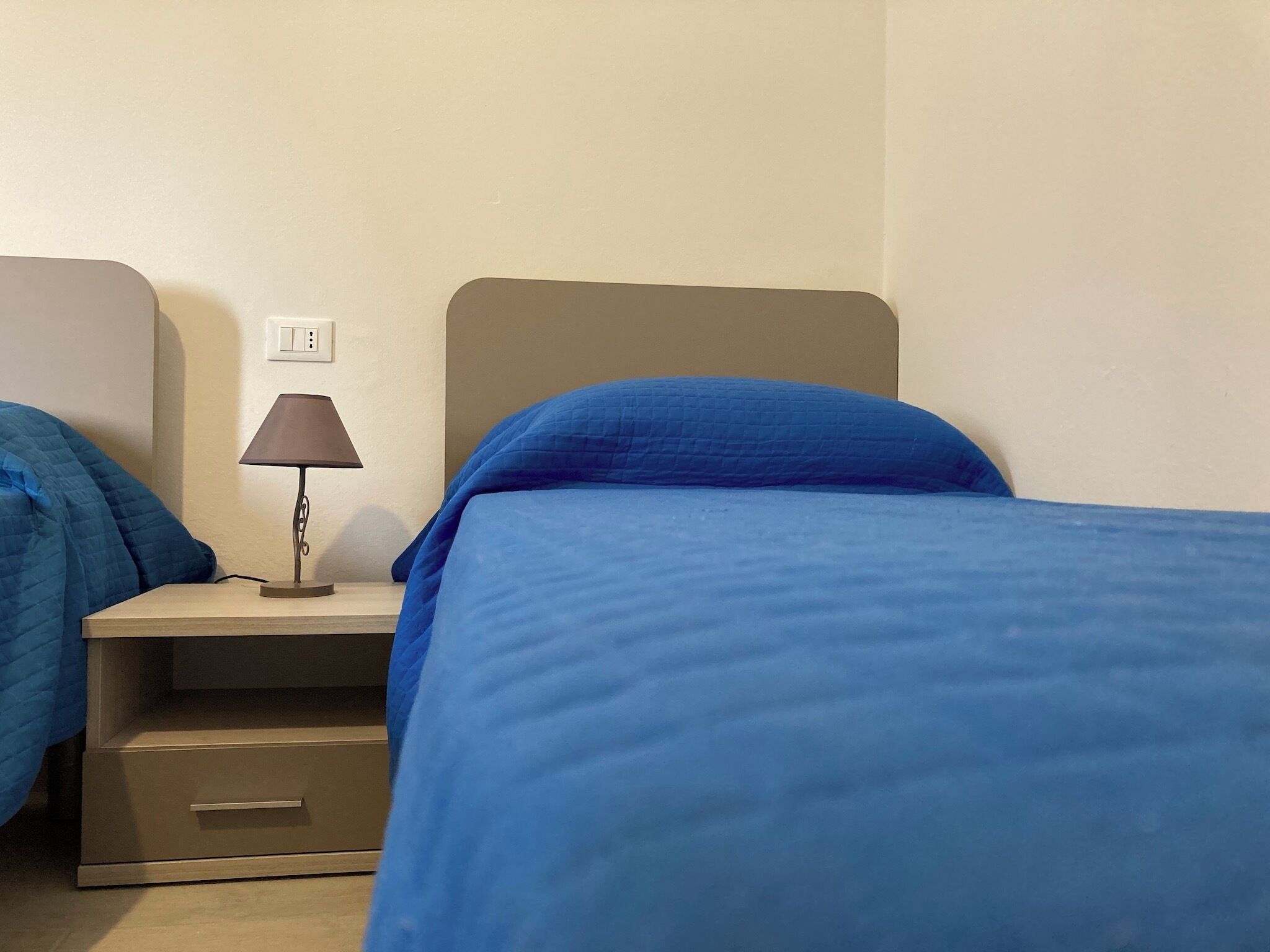 Ein Hotelzimmer mit zwei Betten, jeweils mit blauer Bettdecke und grauem Kopfteil. Ein Nachttisch mit Lampe und Steckdose ist vorhanden.