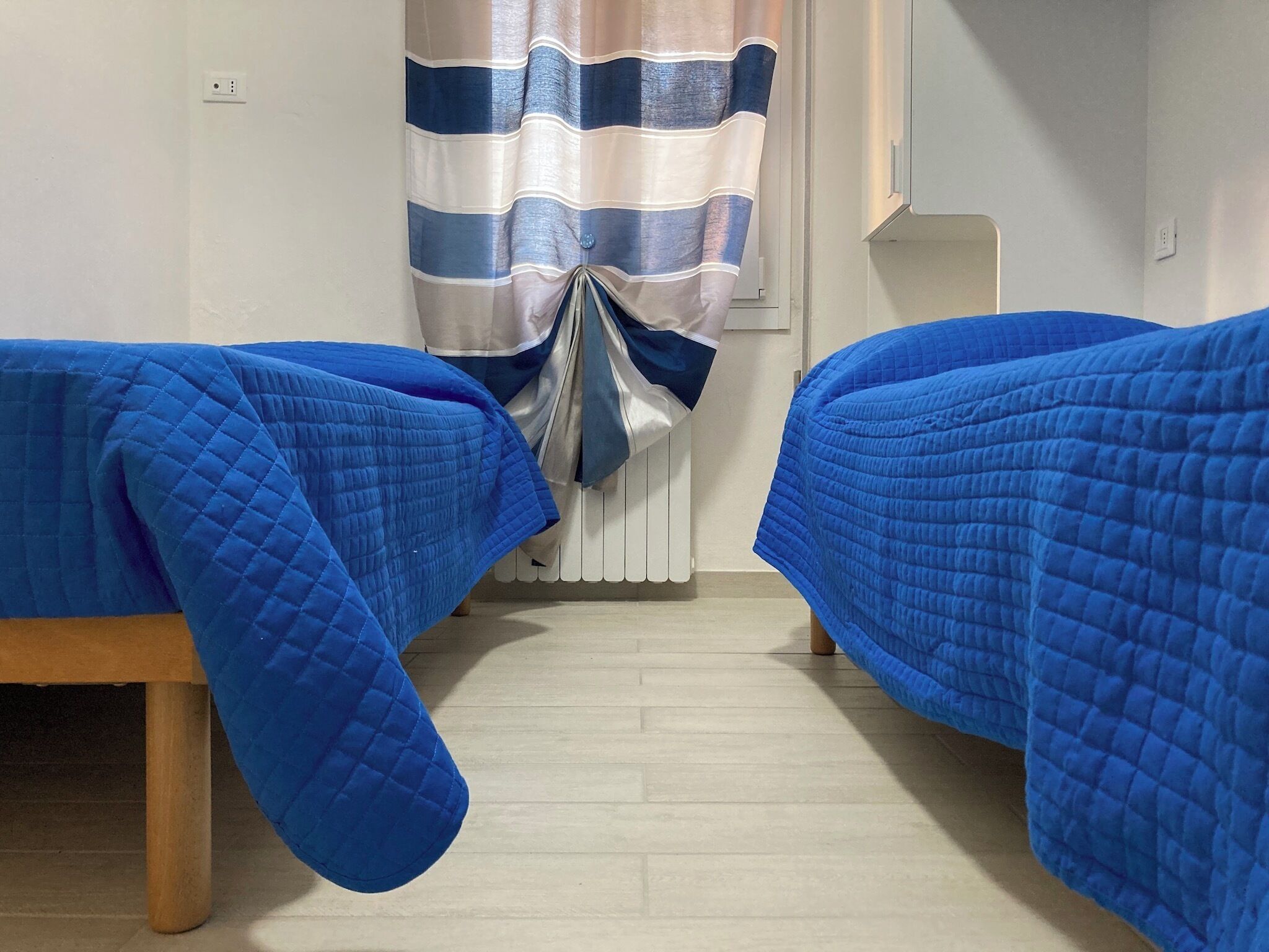 A room with two beds covered in blue quilts, a radiator, and a striped curtain.