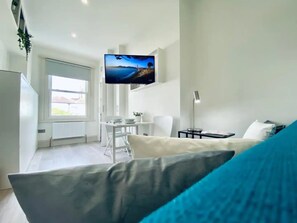 Ein modernes, helles Zimmer mit einem großen an der Wand befestigten Fernseher, einem Essbereich mit Tisch und Stühlen sowie einer gemütlichen Sitzecke mit Sofa und Lampe.