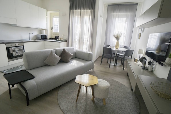 A modern living room with a grey sofa, a small wooden coffee table, a round rug, a dining area with a table and chairs, and a kitchen in the background.