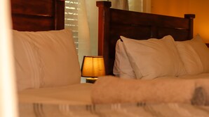 A bed with white bedding and a wooden headboard, a nightstand with a lamp, and a window with blinds.