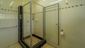 Cabina de ducha con paredes de vidrio, suelo de baldosas, un secador de manos y dispensador de papel higiénico en la pared.