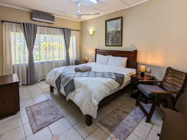Une chambre d’hôtel comprenant un lit, un bureau, une chaise, une table de chevet, un climatiseur, un ventilateur de plafond, une fenêtre avec des rideaux et un tableau au mur.