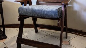 Una silla de madera con cojín azul frente a una pared lisa, sobre suelo de baldosas.