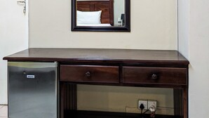 Une pièce avec un bureau en bois, un petit réfrigérateur, un tableau encadré au mur et une porte avec un interrupteur.