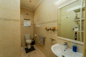 A bathroom with a toilet, sink, mirror, and shelves with toiletries. - Zambezi Family Lodge - Rhino Room (Victoria Falls)