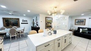 Una cocina amplia con una isla central, armarios blancos y una araña. Hay un comedor con una mesa y sillas, y una sala de estar con un sofá.