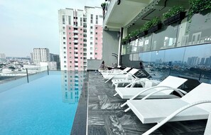 Une piscine à débordement située sur le toit, avec des chaises longues blanches, une vue sur la ville et un gratte-ciel en arrière-plan.