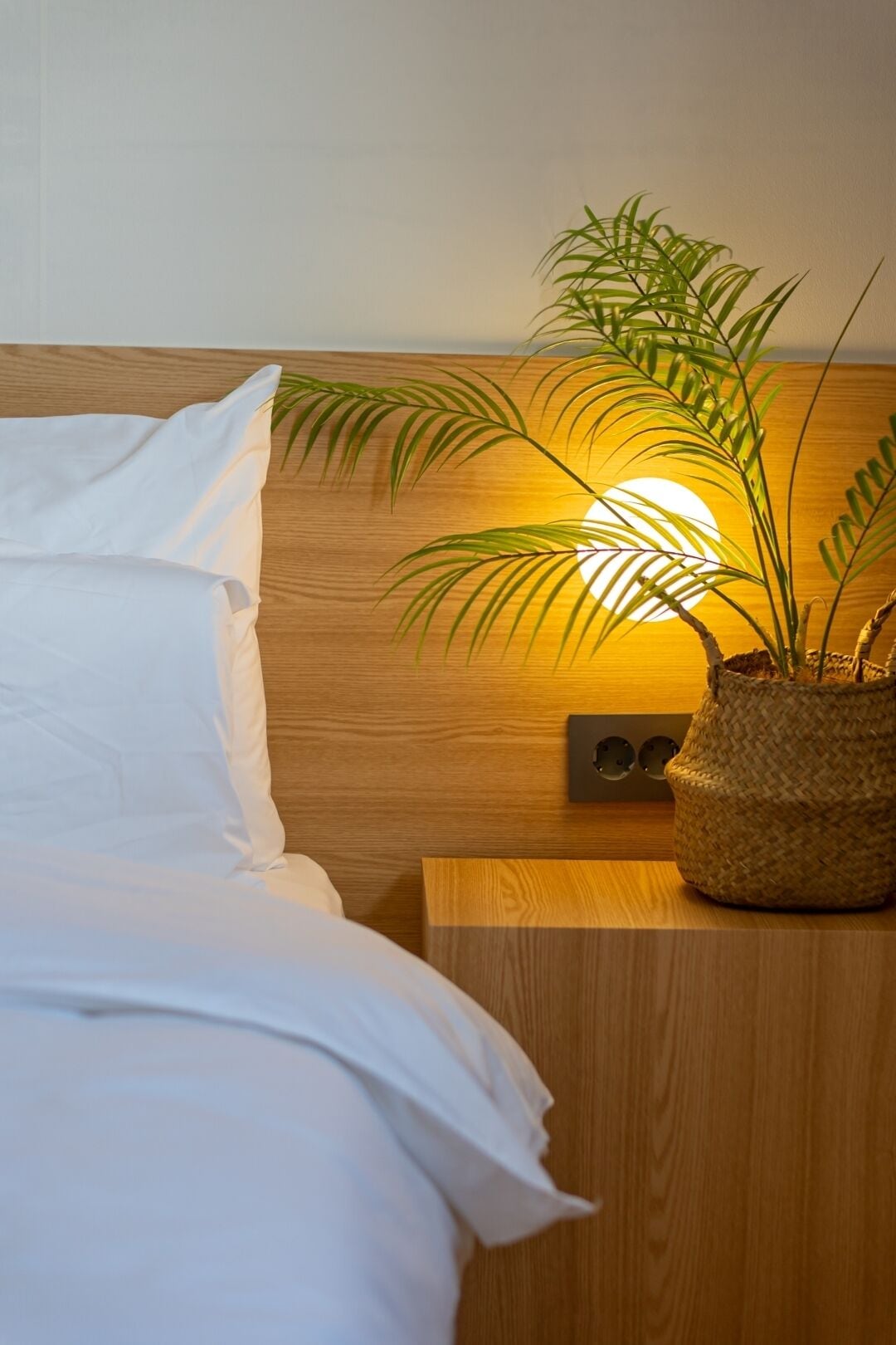 A neatly made bed with white linens, a wooden headboard, a bedside table with a woven basket, and a wall-mounted light fixture.