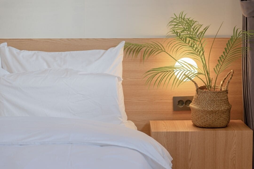 Un lit bien fait, avec du linge de lit blanc, une tête de lit en bois et une table de chevet sur laquelle est posé un pot de fleurs en osier.