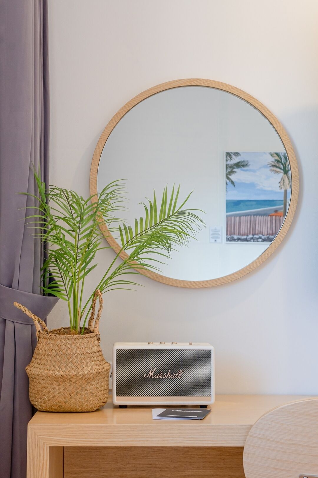 A round mirror with a wooden frame, a woven basket with a plant, and a vintage Marshall radio on a wooden desk.