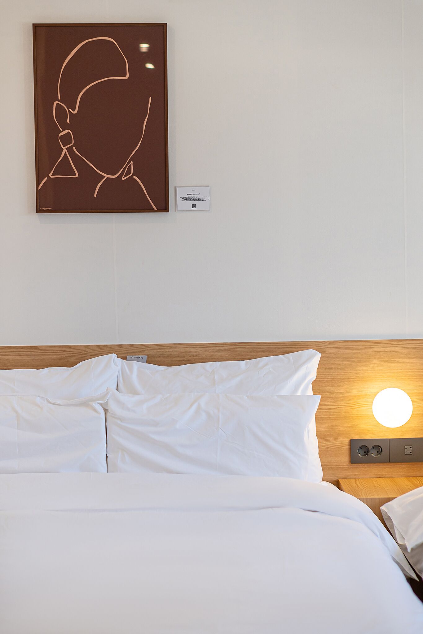 Una cama bien hecha con sábanas blancas y cabecero de madera. Se observa una obra de arte colgada en la pared sobre la cama.