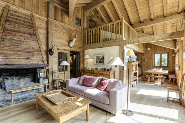 Een rustiek houten interieur met een open haard, een bank, een eettafel en een trap.