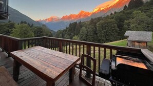 Una terraza de madera con una mesa y sillas, con vistas a un paisaje montañoso donde se distingue una cabaña a lo lejos.