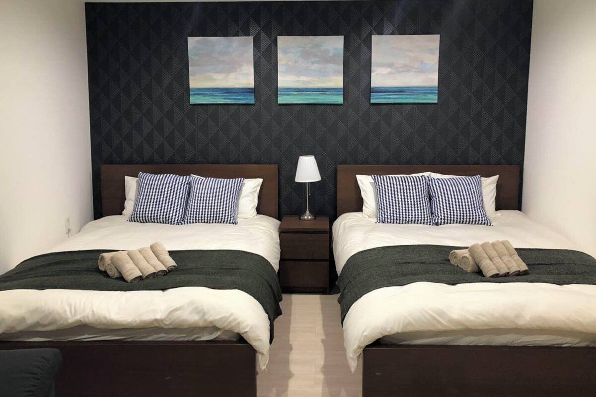 Una habitación de hotel con dos camas, cada una con un cabecero oscuro, ropa de cama blanca y almohadas a cuadros. Hay dos cuadros colgados en la pared que representan una playa.