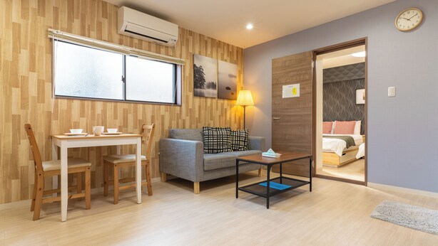 A room with a wooden floor, a gray sofa, a small table, a dining table with chairs, and a bed in the adjacent room. - VALUE NAMBA SOUTH  (Osaka)