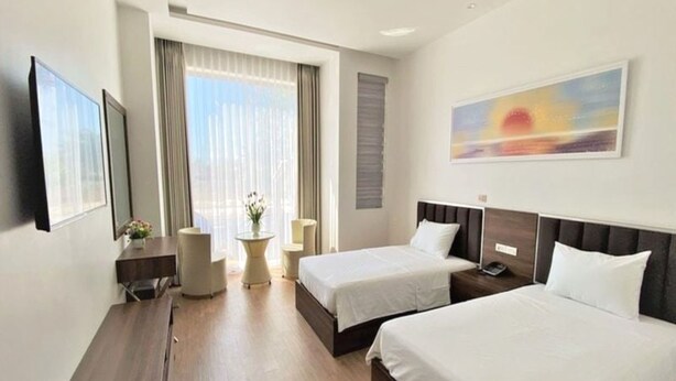 A hotel room with two beds, a desk, a chair, a small table with a vase, and a painting on the wall. - Capital O 1194 Bee Hotel (Nha Trang)