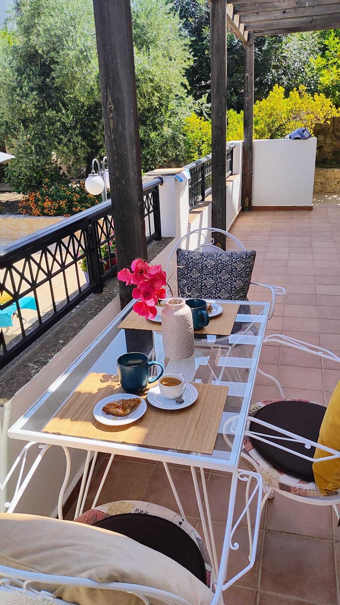 An outdoor patio with white metal chairs and tables, a white wall, and a view of greenery and a pool.