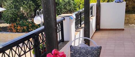 An outdoor patio with white metal chairs and tables, a white wall, and a view of greenery and a pool.