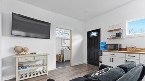 A modern living room with a flat-screen TV, a sofa, and a coffee table. There is a kitchen area with a microwave and a window above the counter. - Hidden Cottage, mins to Main St, Sleeps 4! (Fredericksburg)
