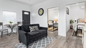 A modern living room with a black sofa, a small table, and a clock on the wall. - Hidden Cottage, mins to Main St, Sleeps 4! (Fredericksburg)
