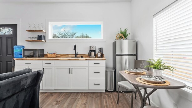 A modern kitchen with white cabinets, a wooden countertop, and a stainless steel refrigerator. There is a window with a view of trees and a framed picture above the sink. - Hidden Cottage, mins to Main St, Sleeps 4! (Fredericksburg)