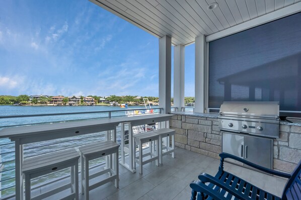 A covered outdoor area with a grill, white tables, and a view of the water and houses in the background. - Blue Heron-Luxury Waterfront Pool Dock (Kingsland)