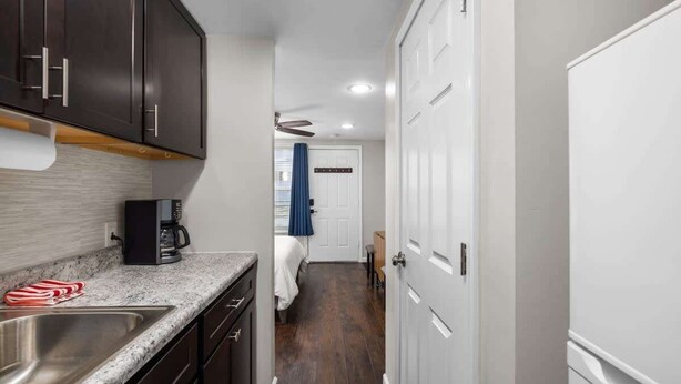 A narrow hallway with a kitchen area on the left, featuring dark cabinets, a stainless steel sink, and a coffee maker. A bed with a blue curtain is visible in the background. - Downtown Kimmswick Studio~Renovated~Prime Location (Imperial)