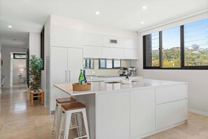 A modern kitchen with white cabinetry, a large island, bar stools, and a view through the window. - Deja Blue by Coast Hosting (Terrigal)