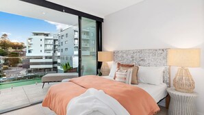 Ein modernes Schlafzimmer mit einem großen Bett, zwei Nachttischlampen und Blick auf eine Stadtkulisse durch eine Glastür.