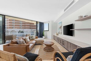 A modern living room with a sofa, coffee table, and wicker chairs, a flat-screen TV, and a large window overlooking a building. - Deja Blue by Coast Hosting (Terrigal)