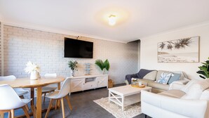 A modern living room with a white brick wall, a flat-screen TV, a dining area with a wooden table and chairs, a white sofa, and a coffee table. - Seadream on Nelson. New listing.
Pet friendly , 300m stroll to beach and cafes.  (Woolgoolga)