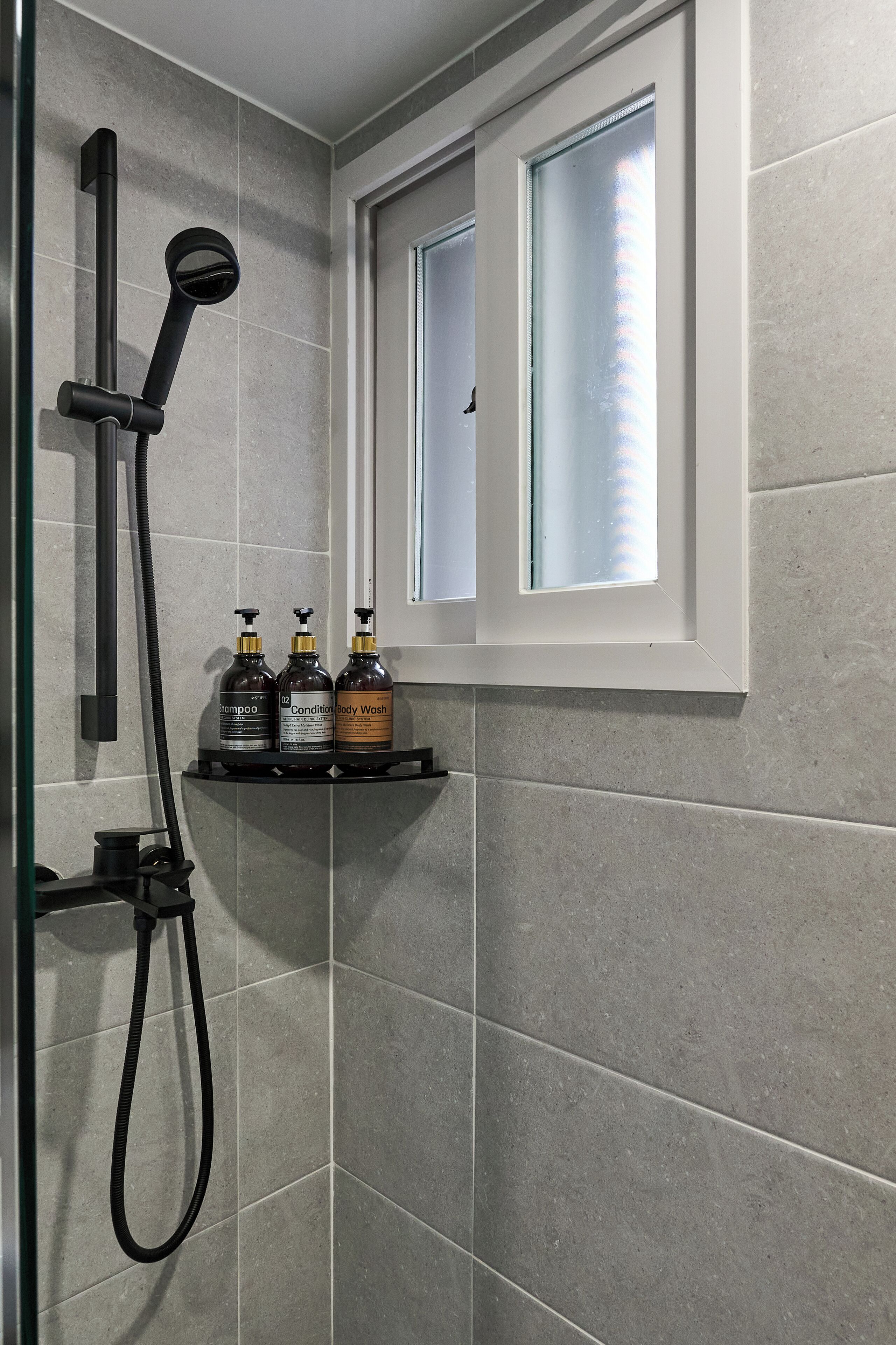 Zona de ducha con azulejos grises, una ventana y tres botellas de champú, acondicionador y gel de baño en un estante.