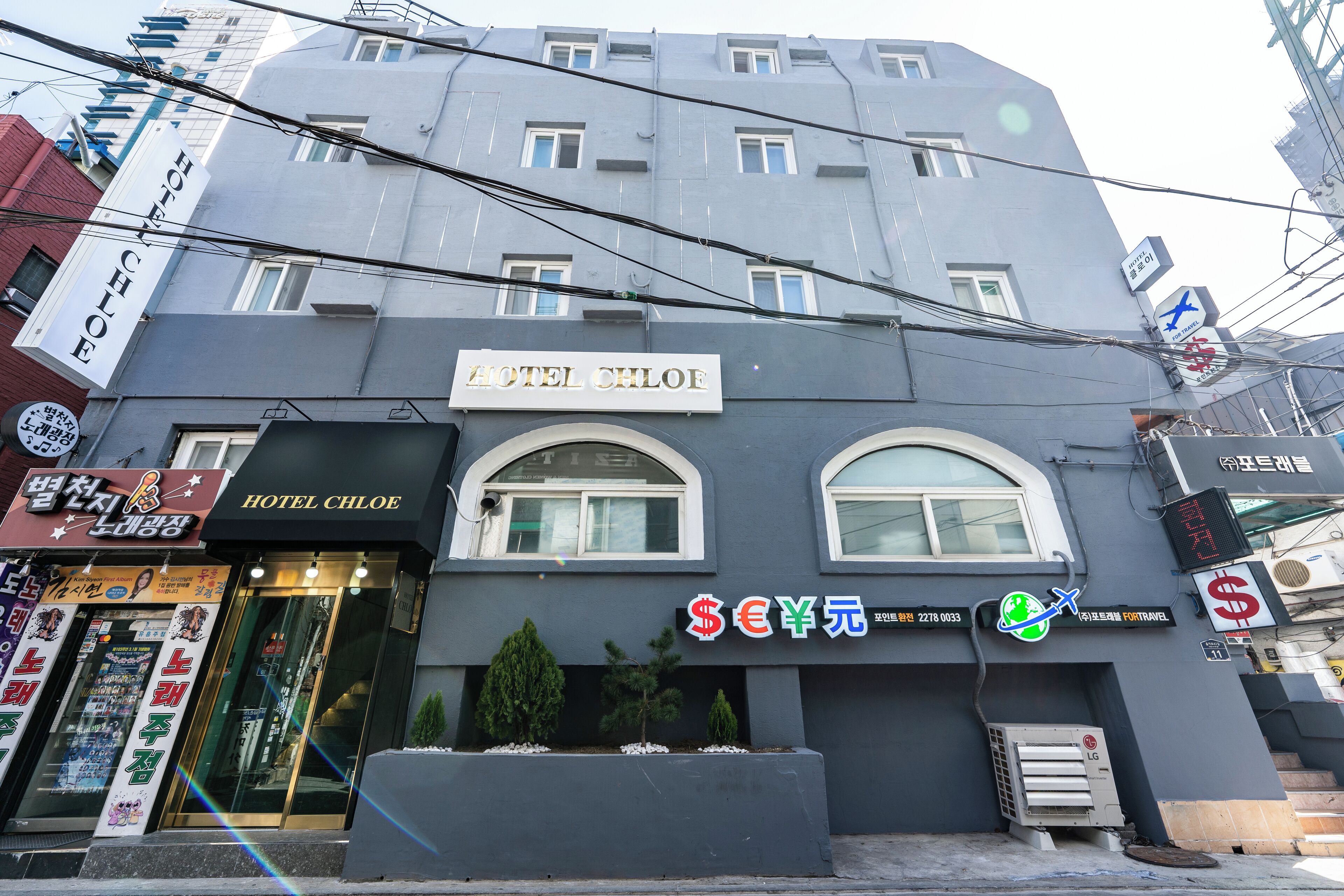 A hotel named 'HOTEL CHLOE' with a signboard, a currency exchange rate display, and a small air conditioning unit in front.