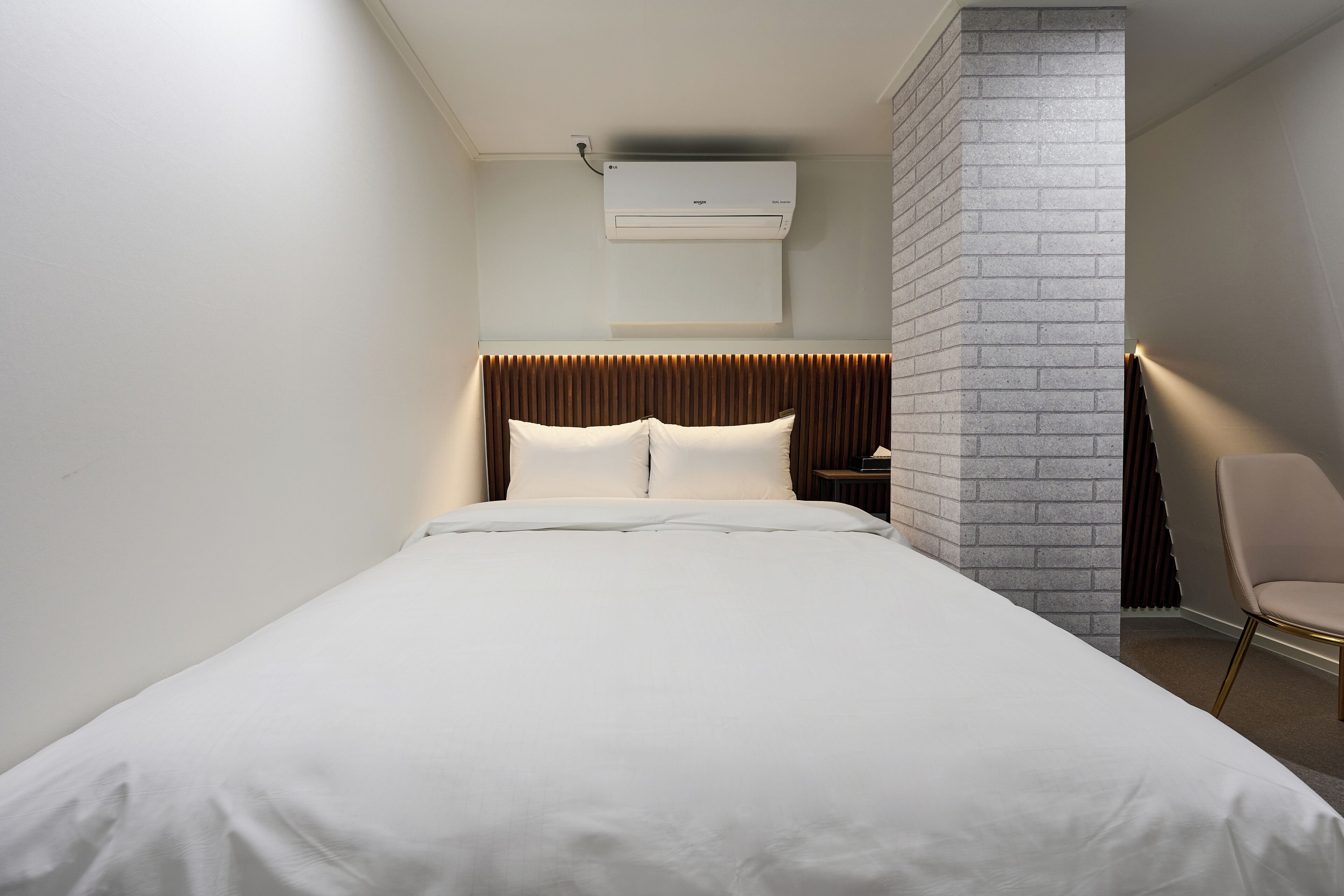 A modern hotel room with a large bed, a brick accent wall, a wall-mounted air conditioner, and a bedside table with a lamp.