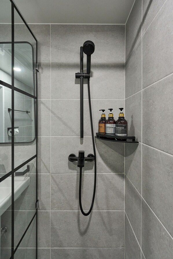 Un baño moderno con baldosas grises, cabina de ducha de vidrio, cabezal de ducha manual negro y un estante con tres artículos de tocador.