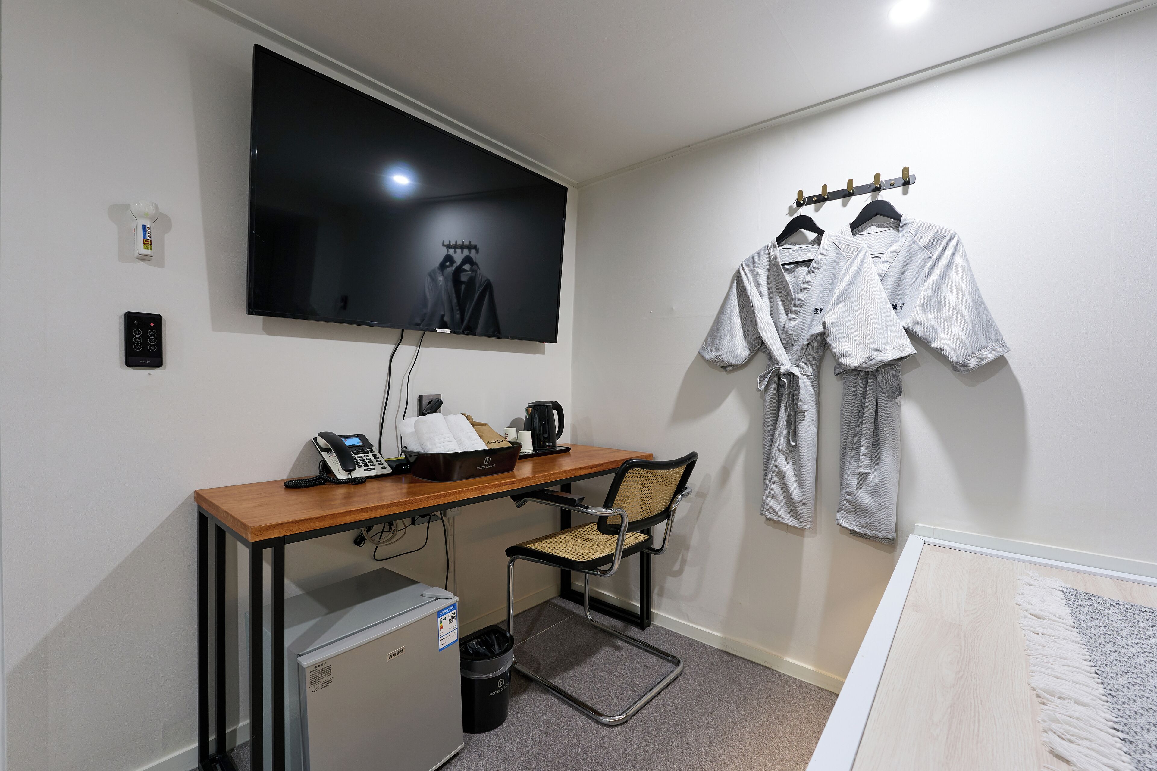 A hotel room with a desk, a TV mounted on the wall, a small refrigerator, a chair, and a clothing rack with robes.